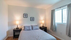 Quarto com cama, duas mesas de cabeceira, duas luminárias de parede, janela com cortinas e duas imagens emolduradas na parede.