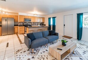 A modern kitchen with wooden cabinets, a stainless steel refrigerator, and a microwave. There is a gray sofa and a wooden coffee table with a remote control.