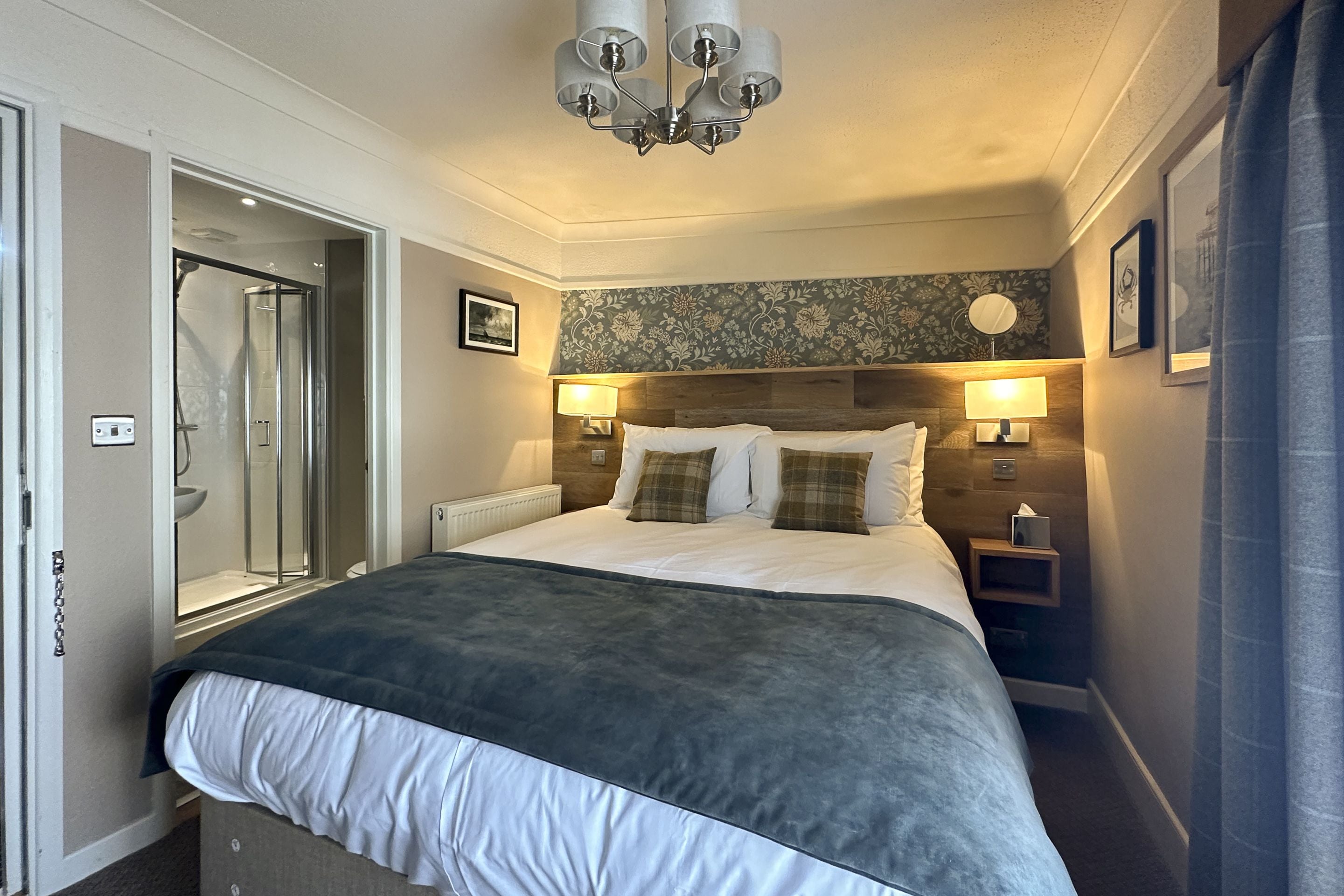 Habitación de hotel con cama, lámparas de noche, mesita de noche, espejo, ducha y puerta.
