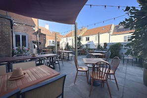 Un patio al aire libre con varias mesas y sillas de madera, rodeado de edificios de ladrillo con tejados de tejas.