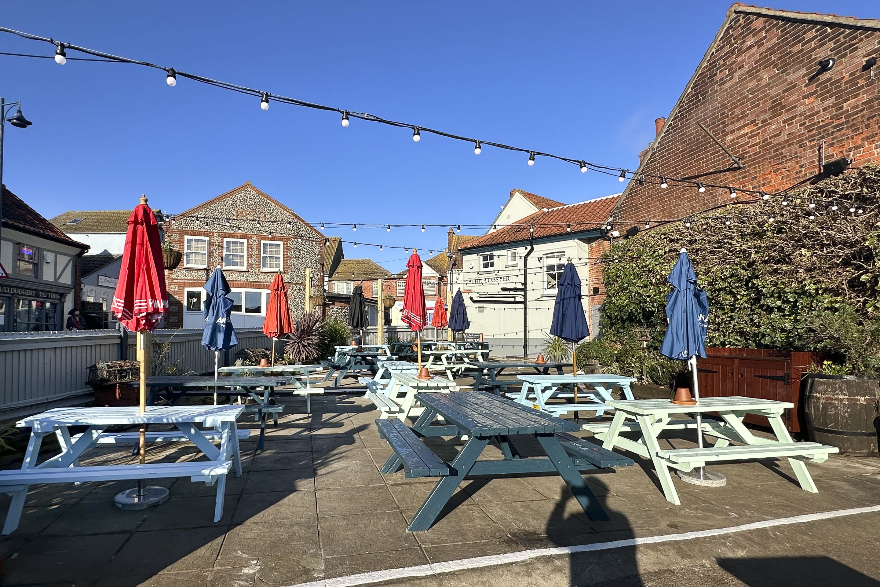 An outdoor seating area with picnic tables and umbrellas, surrounded by buildings.