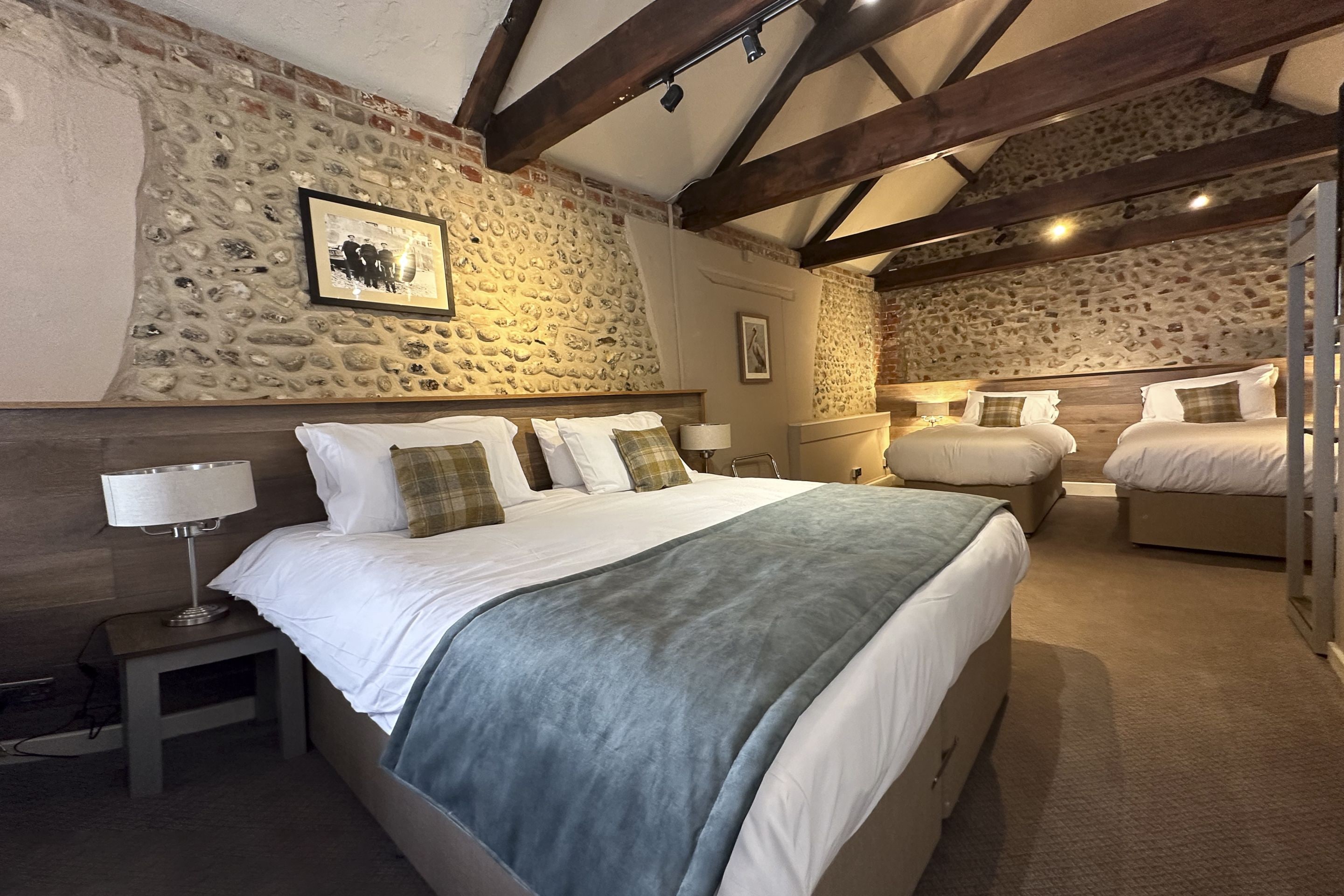 Un dormitorio con pared de piedra, una cama con ropa blanca, una mesita de noche con lámpara y techo de madera.