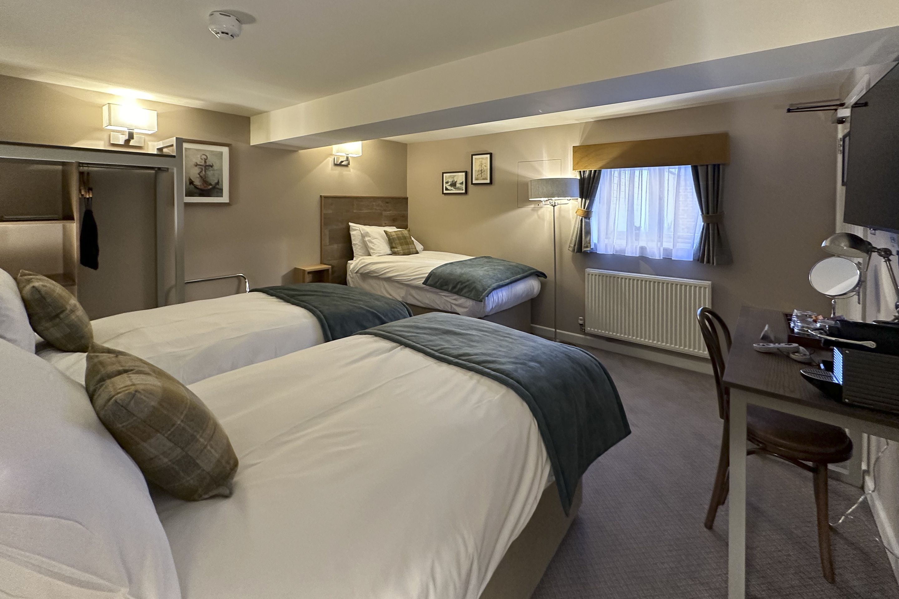 A hotel room with two beds, a desk with a chair, a radiator, and a window with curtains.