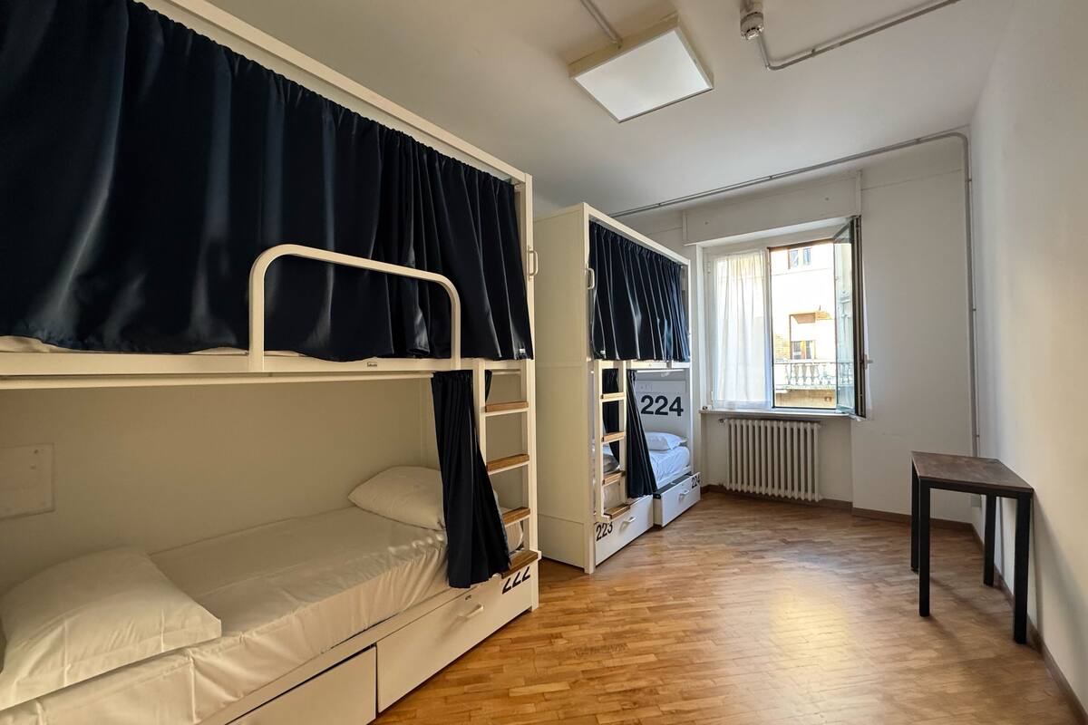 A dormitory room with bunk beds, a small table, and a window with curtains.