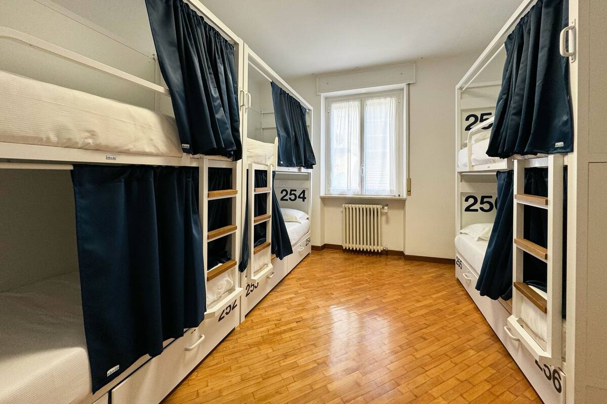 A dormitory room with bunk beds, numbered storage compartments, a window with curtains, and a radiator.
