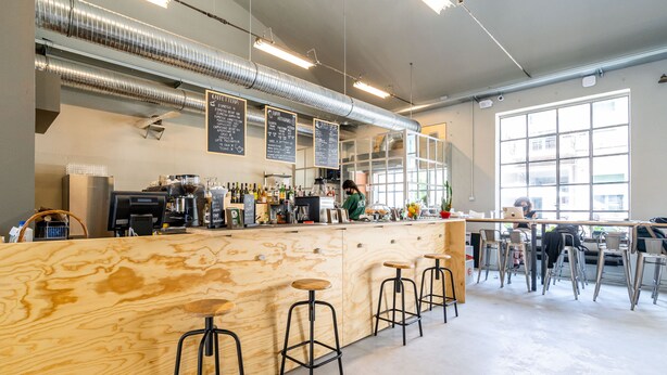 Una cafetería moderna con mostrador de madera, taburetes y un panel con la carta.
