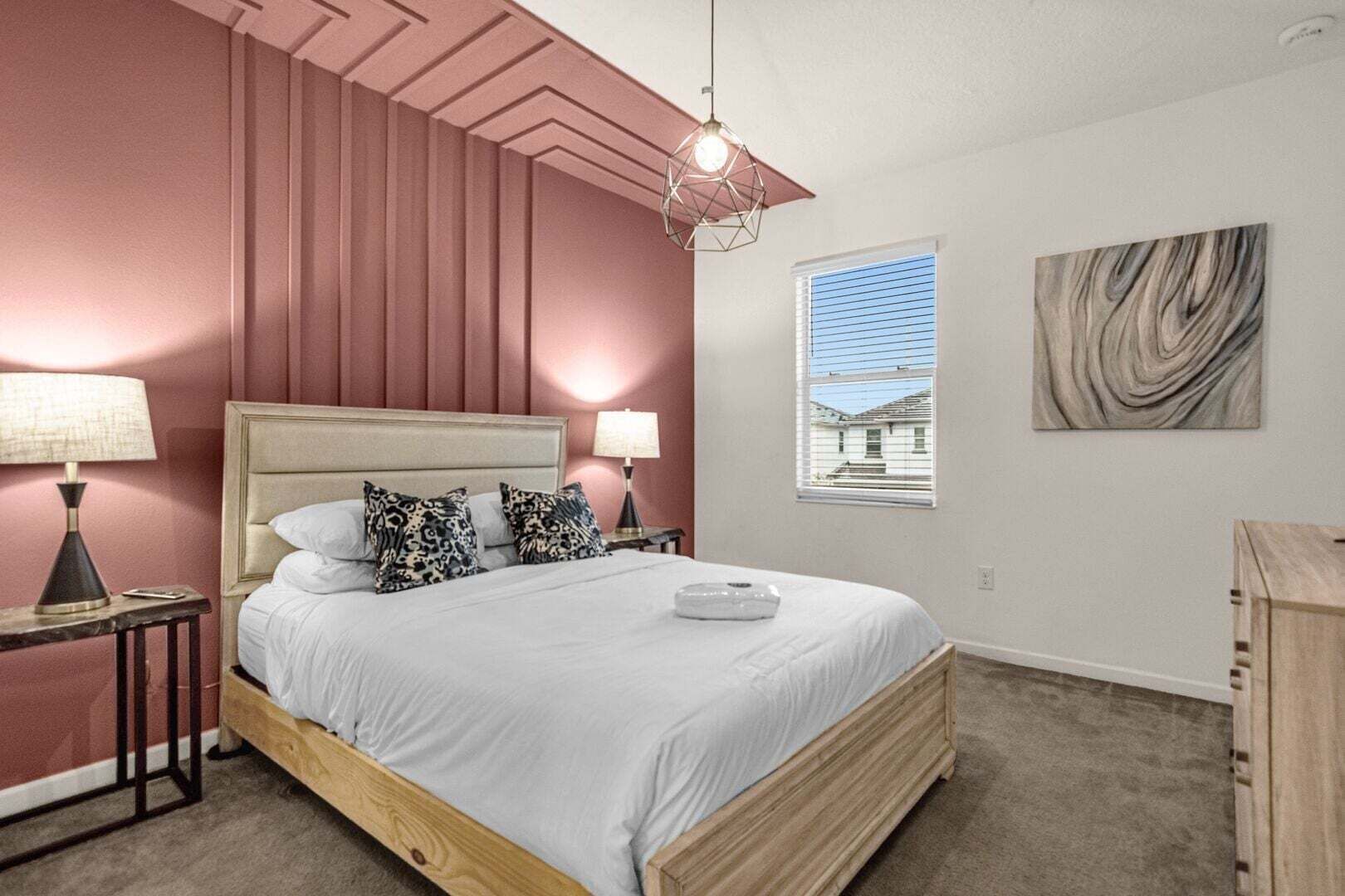A bedroom with a bed, two bedside lamps, a window with blinds, and a wall-mounted artwork.
