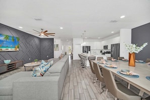Uma cozinha moderna com ilha central, área de jantar com mesa comprida e sala de estar com sofá e TV fixada na parede.