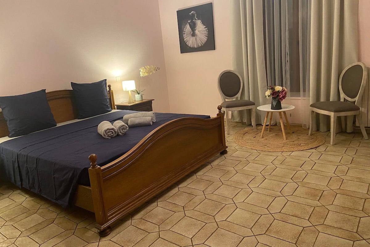 A hotel room with a wooden bed, a small round table with a vase, two chairs, a nightstand with a lamp, and a wall-mounted picture.