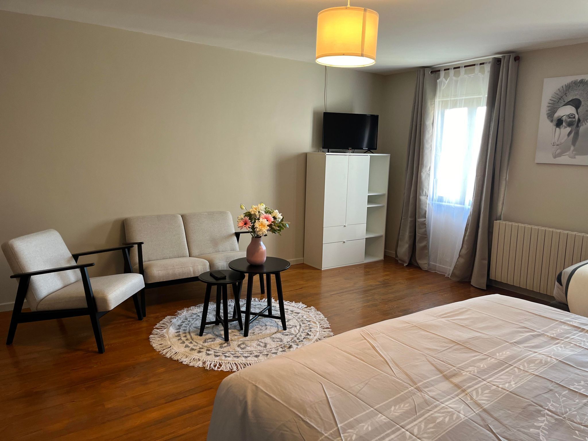 A hotel room with a bed, a sofa, a chair, a small table with a vase of flowers, a TV on a cabinet, and a window with curtains.