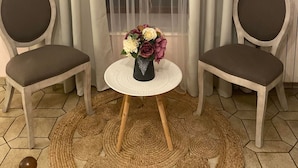 A room with two chairs, a small round table with a vase of flowers, and a window with sheer curtains.