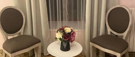 A room with two chairs, a small round table with a vase of flowers, and a window with sheer curtains.