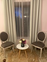 Una habitación con dos sillas, una mesita redonda con un jarrón de flores y una ventana con cortinas ligeras.