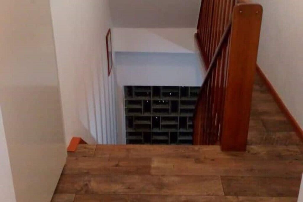 Una escalera de madera que lleva a un rellano con una pared blanca y una rejilla de ventilación.