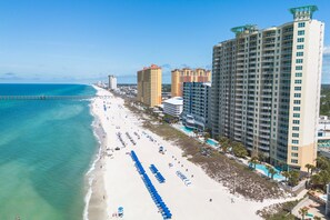A coastal hotel with multiple high-rise buildings, a beach with umbrellas, and a pier extending into the ocean.