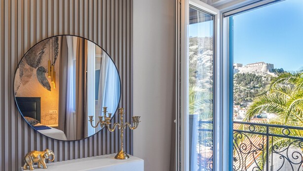 Un baño moderno con un espejo grande, un tocador blanco, un otoman azul y un balcón con vistas a una colina.