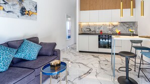 Una sala de estar moderna con un sofá gris, una mesa de centro azul y una cocina con encimera de mármol y luces colgantes.