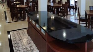 Uma sala de jantar ampla, com mesas e cadeiras de madeira, uma ilha central com bancada preta e iluminação pendente.