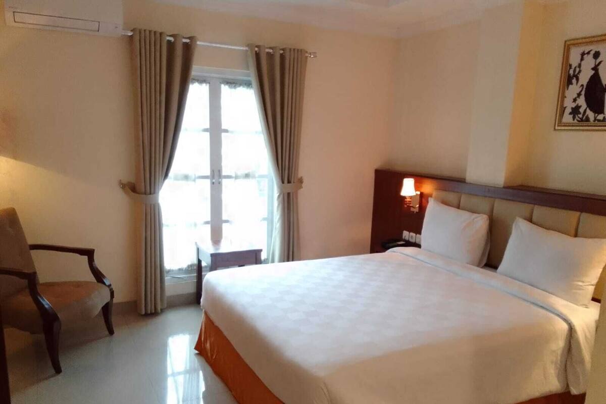 A hotel room with a large bed, a chair, a nightstand with a lamp, and a window with curtains.