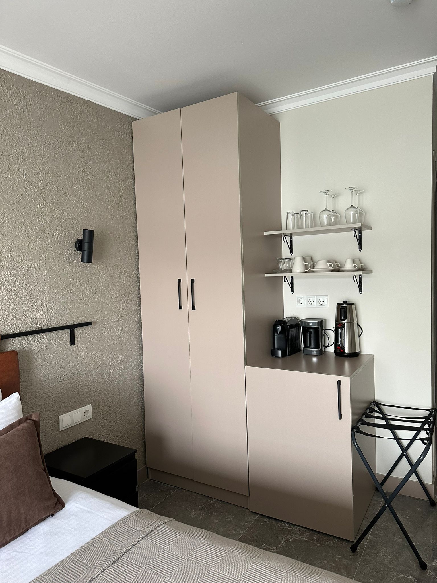 Habitación pequeña con cama, armario, una mesita con tetera y estantería con cristalería.