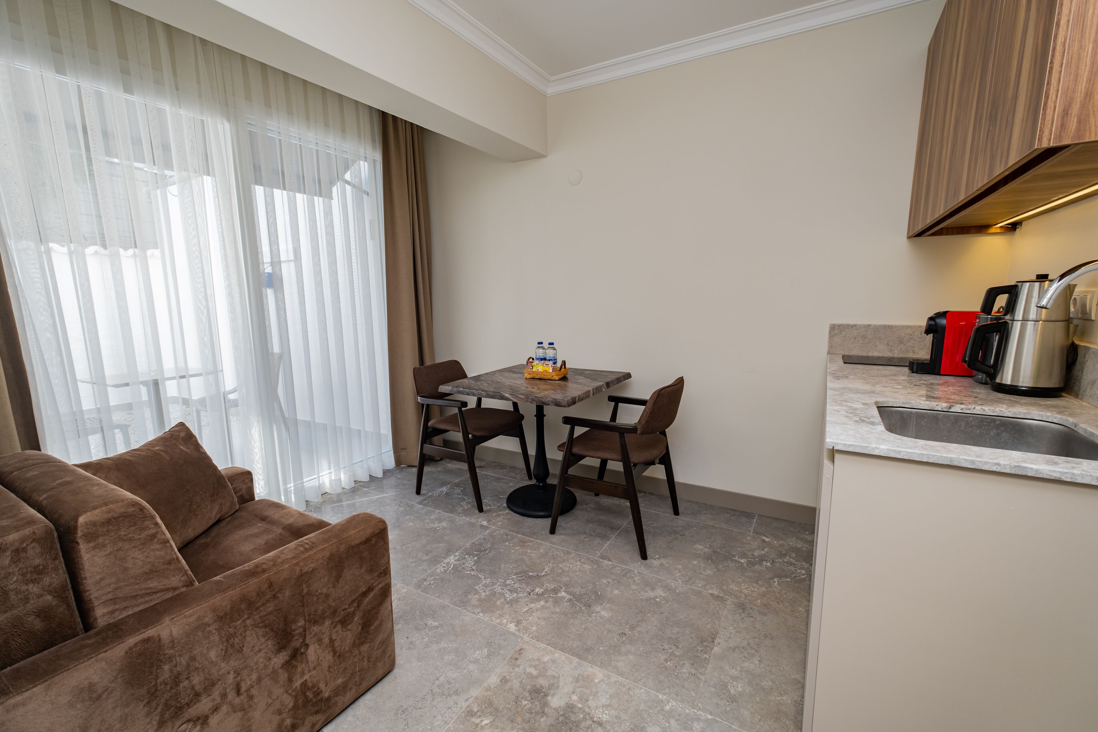 A compact living space with a sofa, a dining table with chairs, a kitchen area with a sink and coffee maker, and a window with sheer curtains.