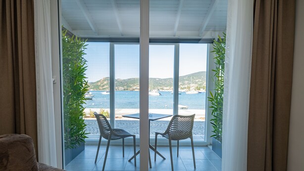 A room with a sliding glass door leading to a balcony with two chairs and a table, overlooking a coastal area with boats and hills.