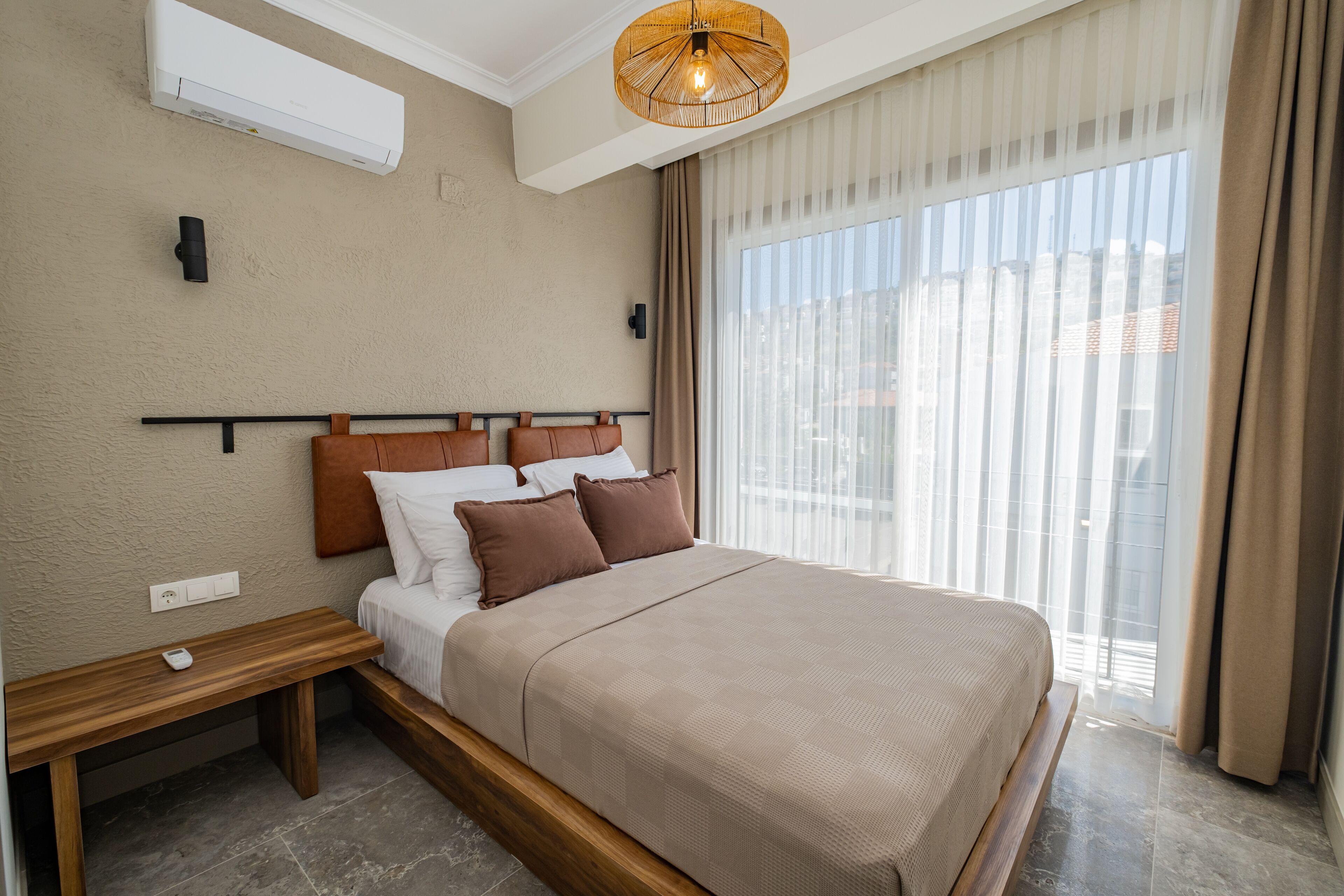 A modern hotel room with a large bed, a bedside table, a wall-mounted air conditioner, and a large window with curtains.