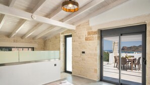 A modern bedroom with a large sliding glass door leading to a balcony with a table and chairs, stone walls, and a view of the sea. - Villa Megalith (Chania)