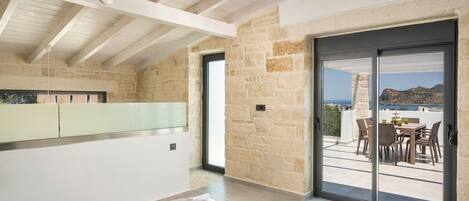 A modern bedroom with a large sliding glass door leading to a balcony with a table and chairs, stone walls, and a view of the sea.