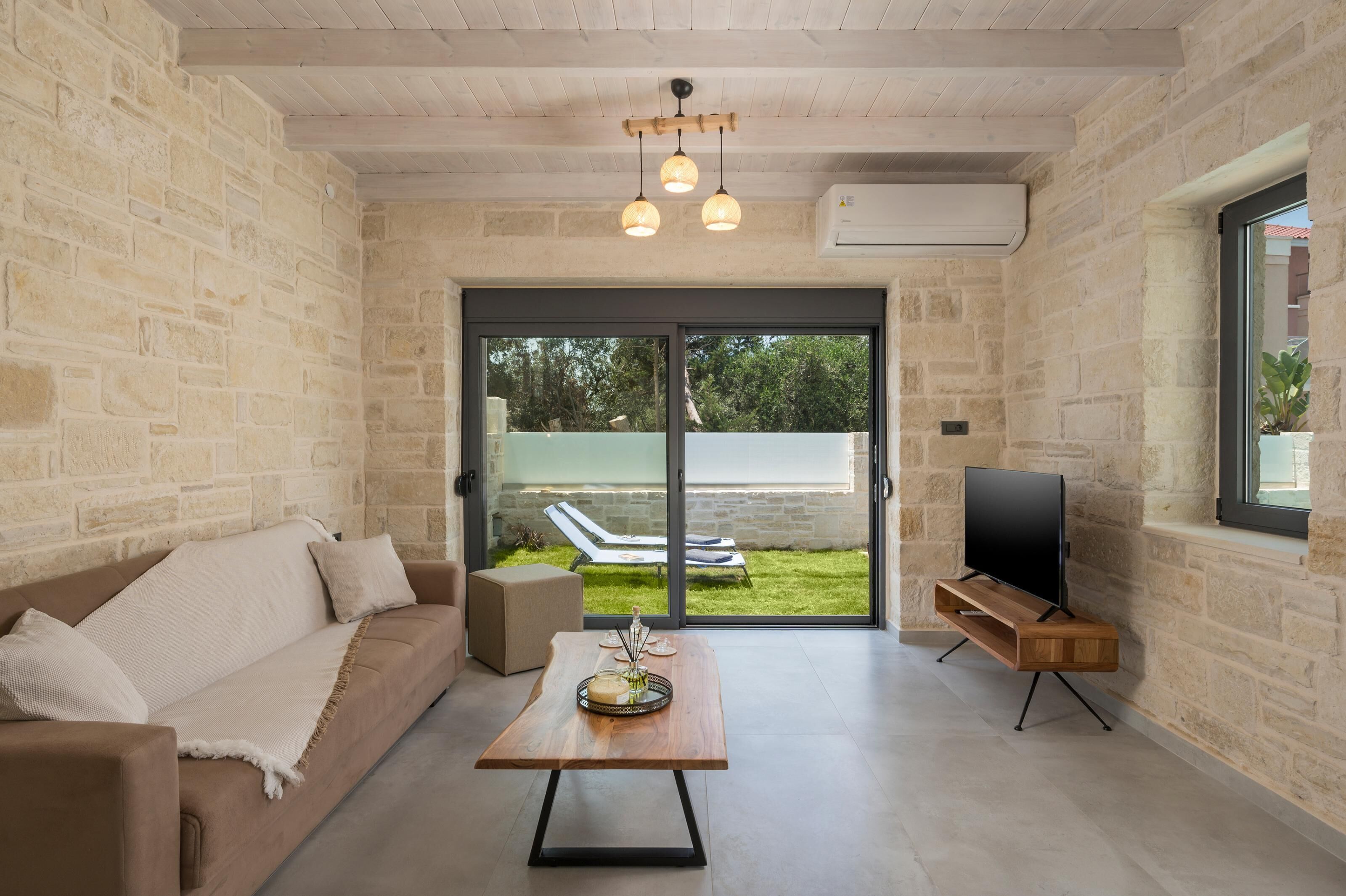 A modern living room with a stone wall, a sofa, a coffee table, a TV on a wooden stand, and a sliding glass door leading to a garden.