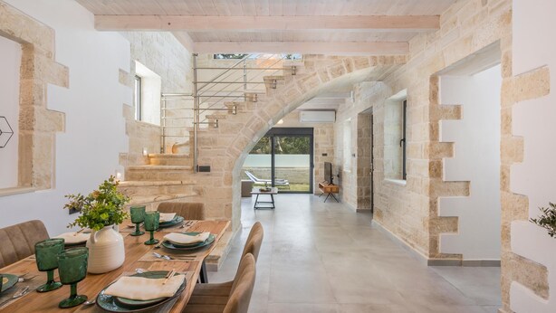 A spacious dining area with a high stone wall, a staircase leading to a balcony, and a view of the outdoors. - Villa Megalith (Chania)
