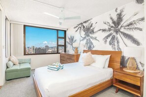 A hotel room with a large window, a bed with white linens, a green sofa, wooden nightstands, and a cityscape view. - Ocean-view condo steps to Waikiki Beach (Honolulu)