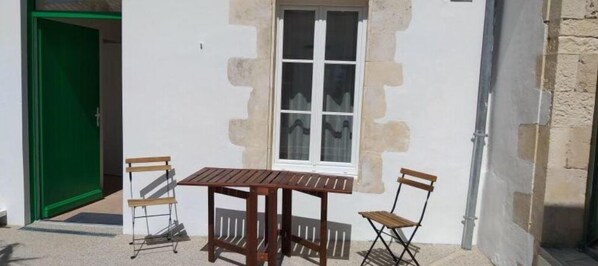 Ein kleiner Sitzbereich im Freien mit einem Holztisch und zwei Stühlen, einem Fenster mit weißem Rahmen und einer grünen Tür.