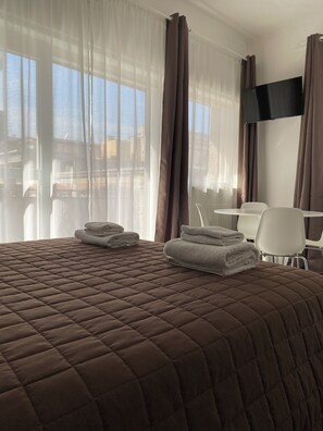 A hotel room with a large bed, a flat-screen TV mounted on the wall, and a view of the cityscape through the windows. - Quintino Sella rooms (Bari)
