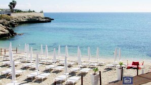 Une plage bordée de rangées de transats et de parasols blancs, un littoral rocheux et une vue dégagée sur la mer.