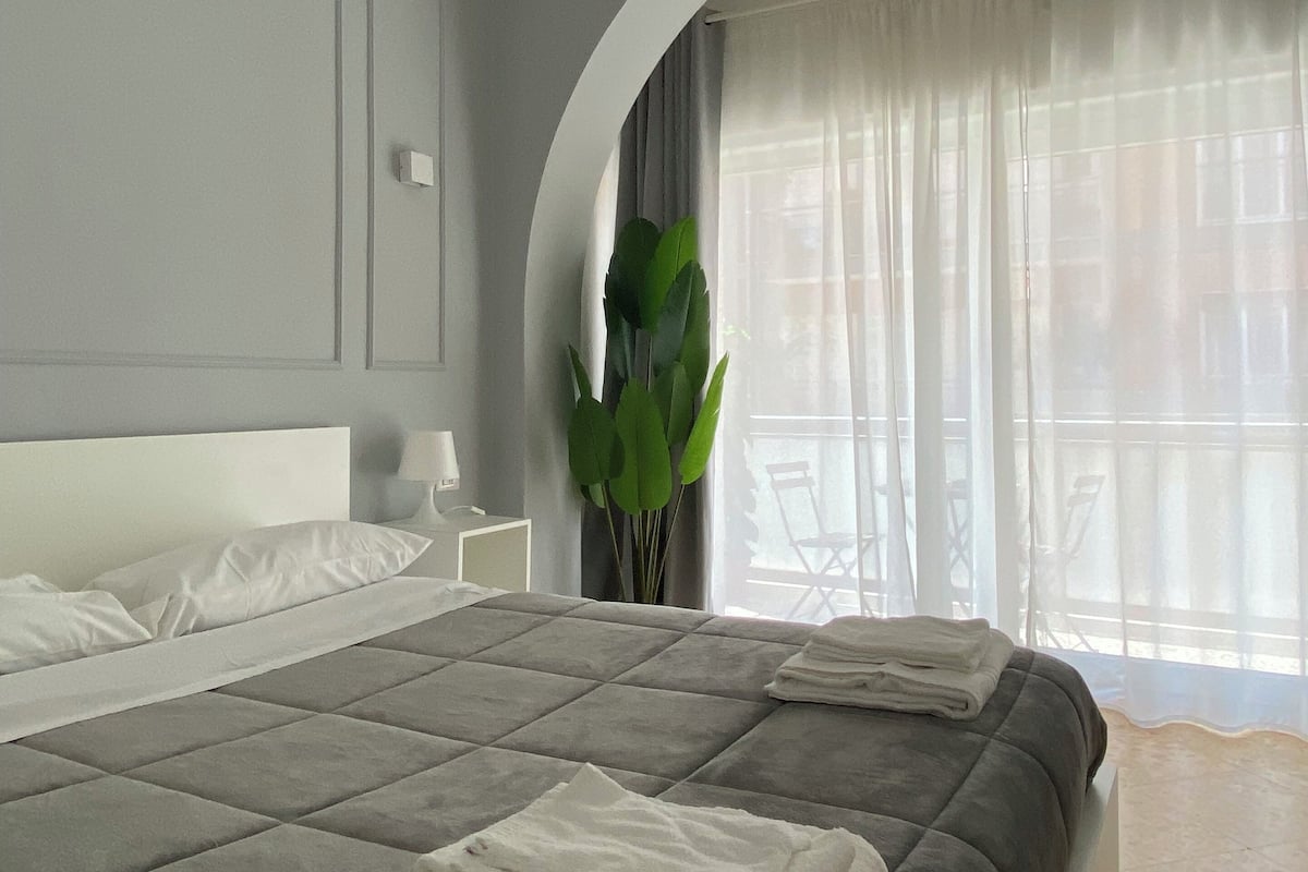 A modern bedroom with a large bed, a bedside table, a plant, and a view through a window with curtains.