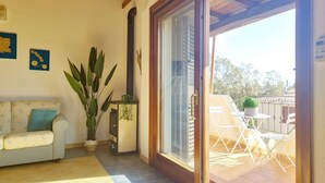 En hyggelig stue med en sofa, en stor plante og en skydedør, der fører ud til en udendørs terrasse med stole og et bord.