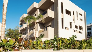 A modern multi-story building with balconies and a flat roof, surrounded by tropical plants.