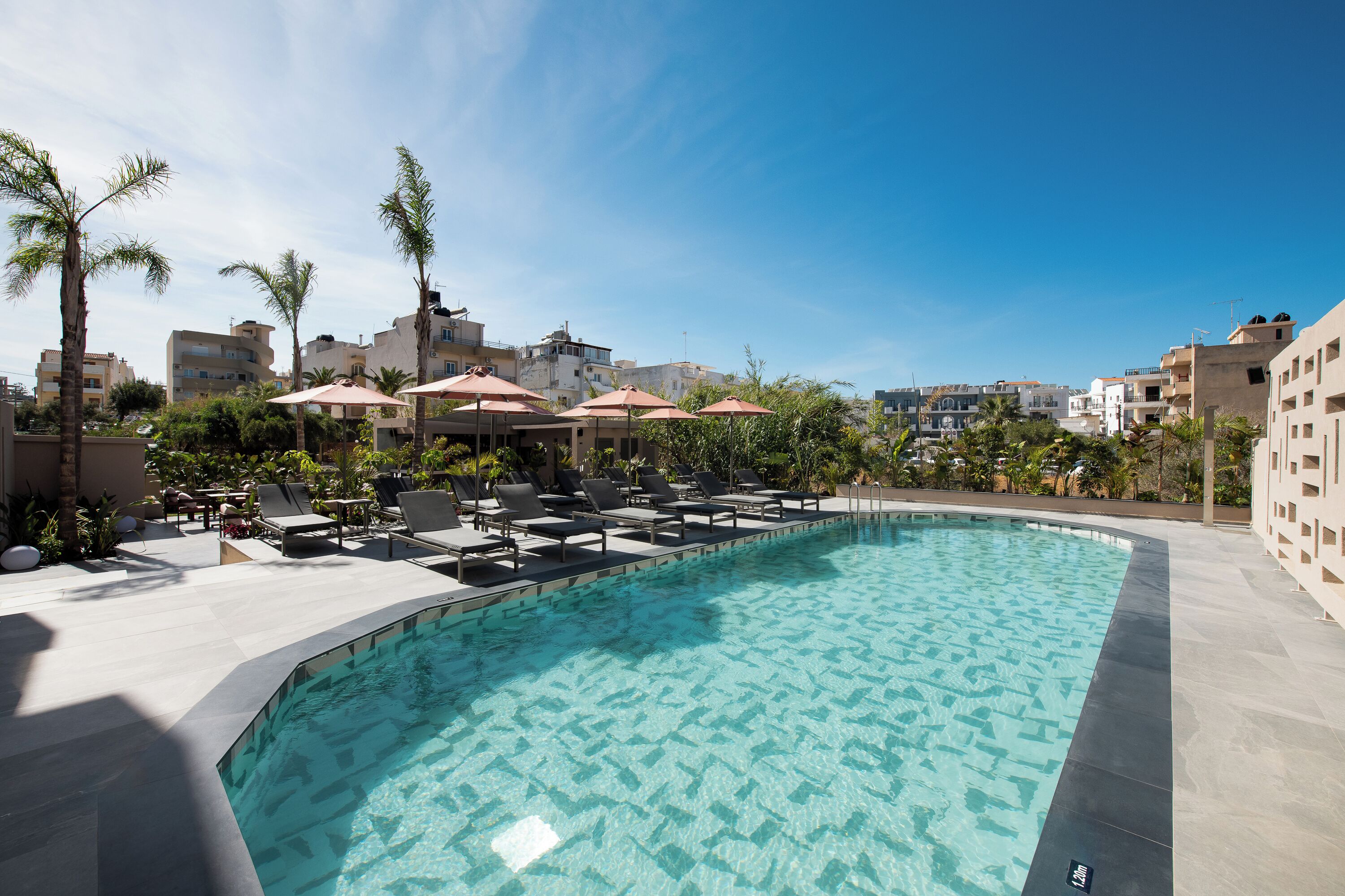 A swimming pool with lounge chairs and umbrellas, surrounded by buildings and palm trees.