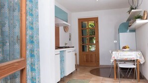 A compact kitchen with a wooden door, white cabinets, a small table with a bowl of fruit, and a patterned curtain. - [Sunrise] Seaview apartment with large terrace (Orosei)