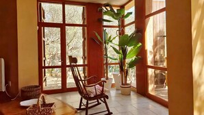 Una habitación con ventanas amplias, un sillón mecedor, una planta en maceta y una cesta tejida.