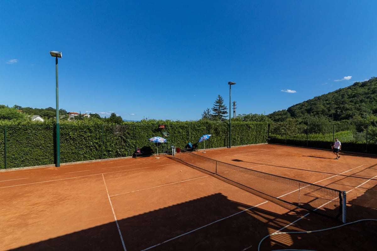 An outdoor tennis court with a net, surrounded by greenery and a clear sky.