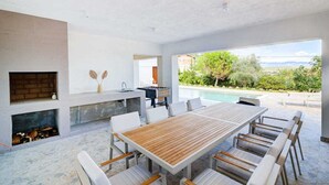 A modern outdoor dining area with a long wooden table and white chairs, a built-in grill, and a view of a pool and greenery. - Villa Dreamview by Aneo Travel (Matulji)