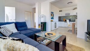 A modern living room with a sofa, coffee table, and a TV. There is a kitchen in the background with white cabinets and a stove.