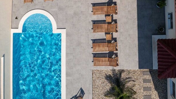 An aerial view of a pool area with a large pool, wooden lounge chairs, and a red-roofed structure.
