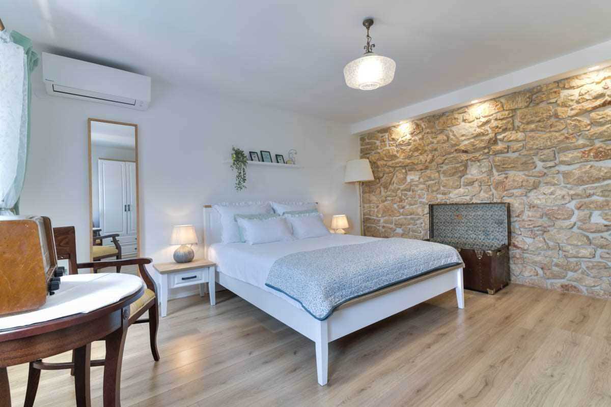 A bedroom with a stone wall, a bed with a white frame, a bedside table with a lamp, and a wooden chair.