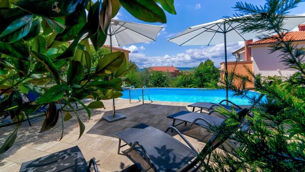Zona de piscina con sillones de descanso y sombrillas, rodeada de vegetación y edificios al fondo.
