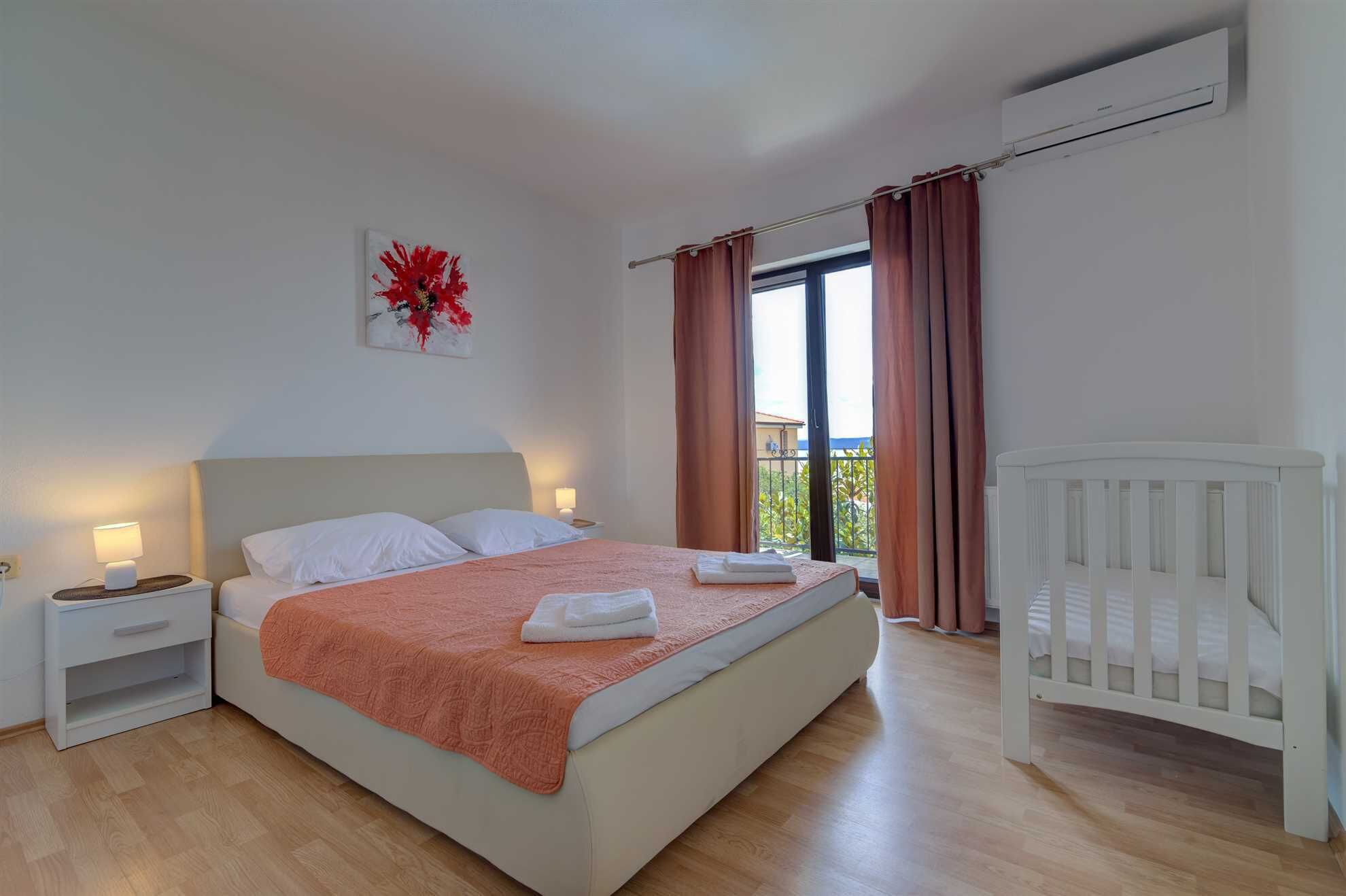 A bedroom with a bed, nightstands, a crib, a window with curtains, and an air conditioning unit.