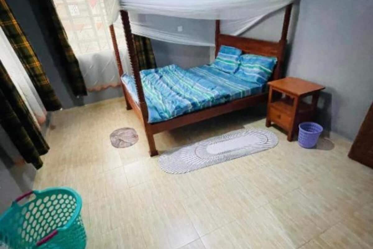 A bedroom with a bed, a small wooden nightstand, a mat, a basket, and curtains.