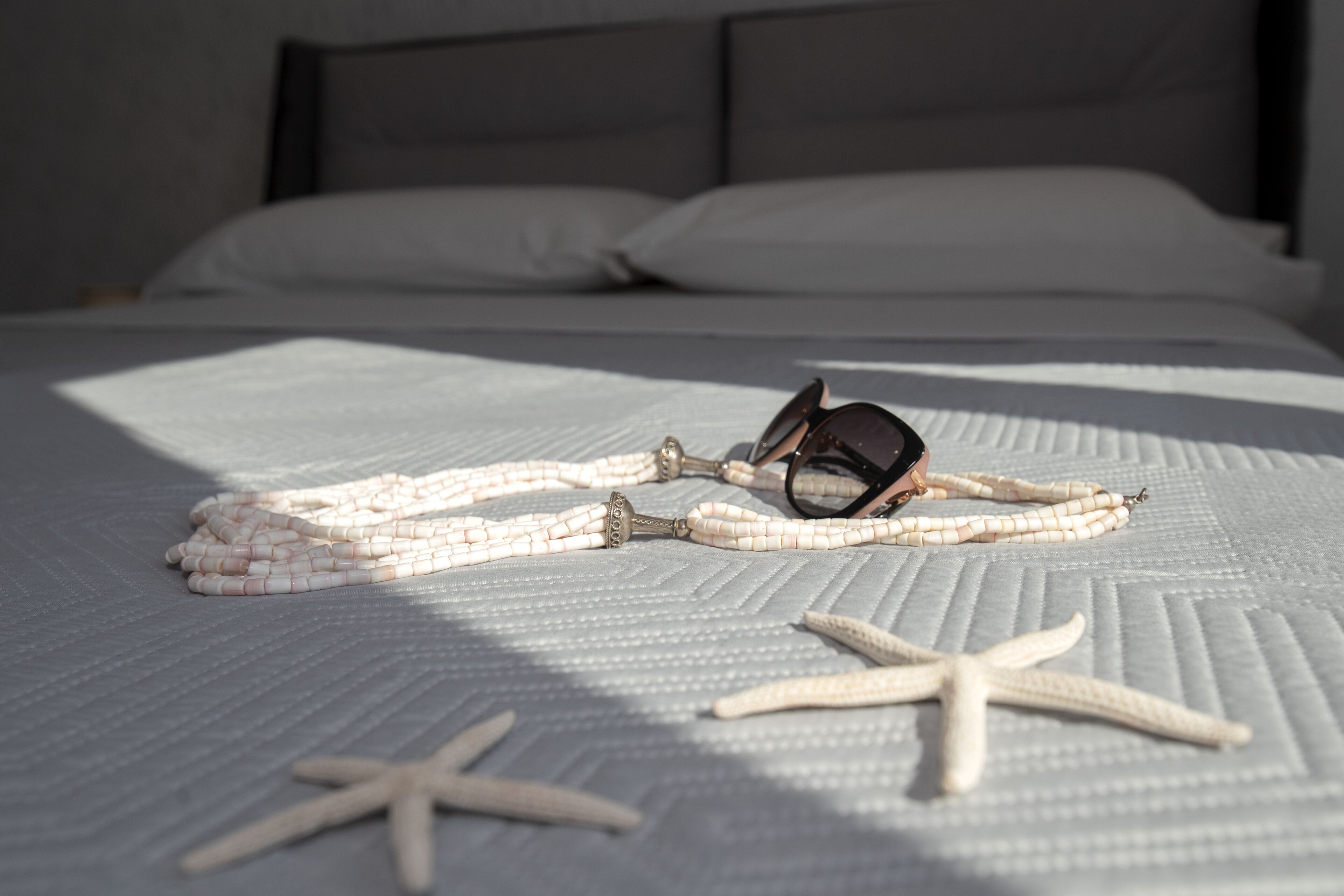 A bed with a white sheet, a pair of sunglasses, a beaded necklace, and a starfish.